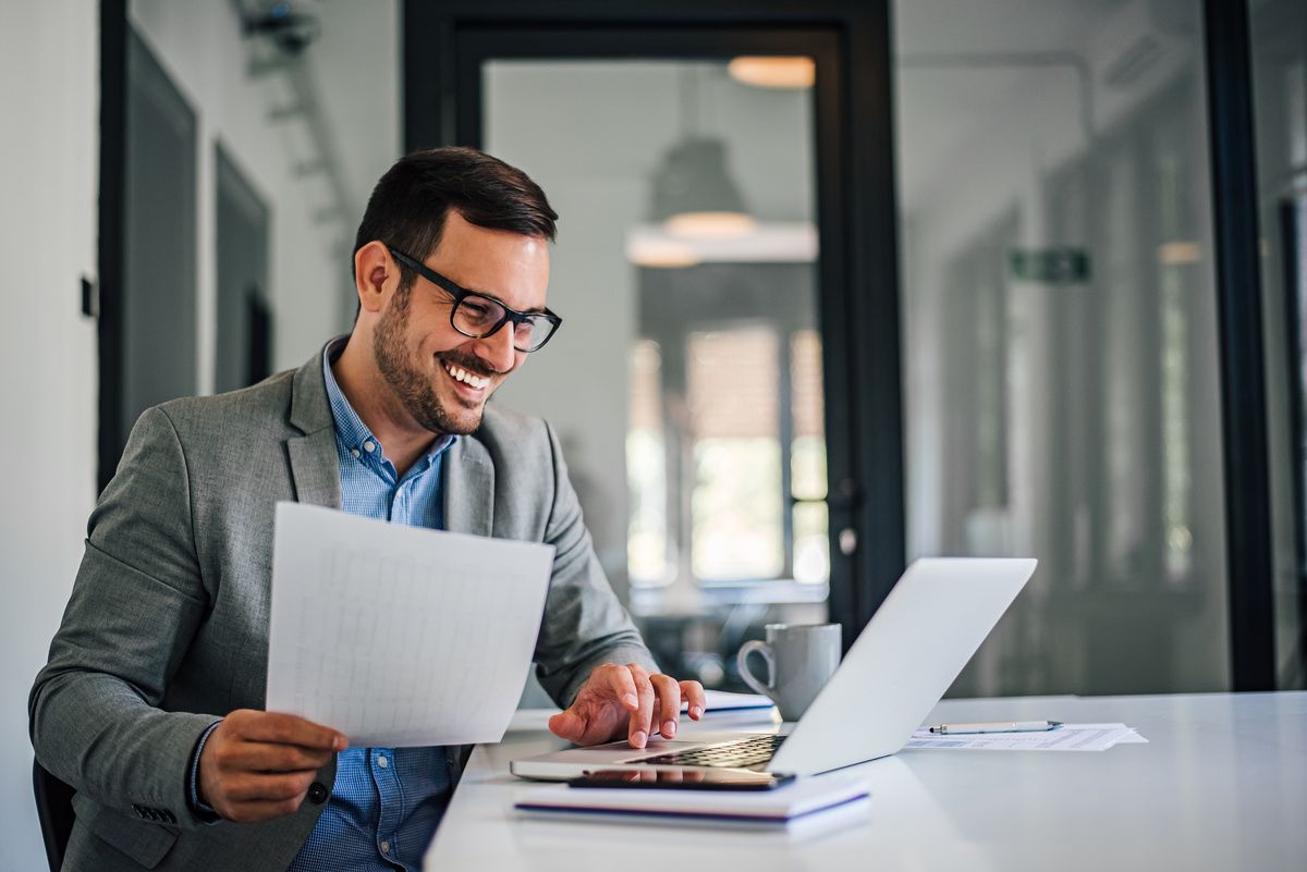 Portrait of young smiling cheerful entrepreneur businessman in bright modern office working with laptop computer examining charts, graphs and paper documents